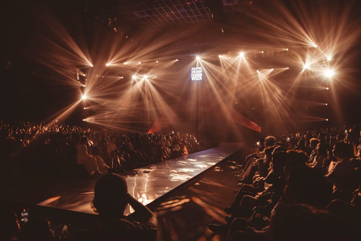 Models walking a runway under catwalk lighting at a fashion show.