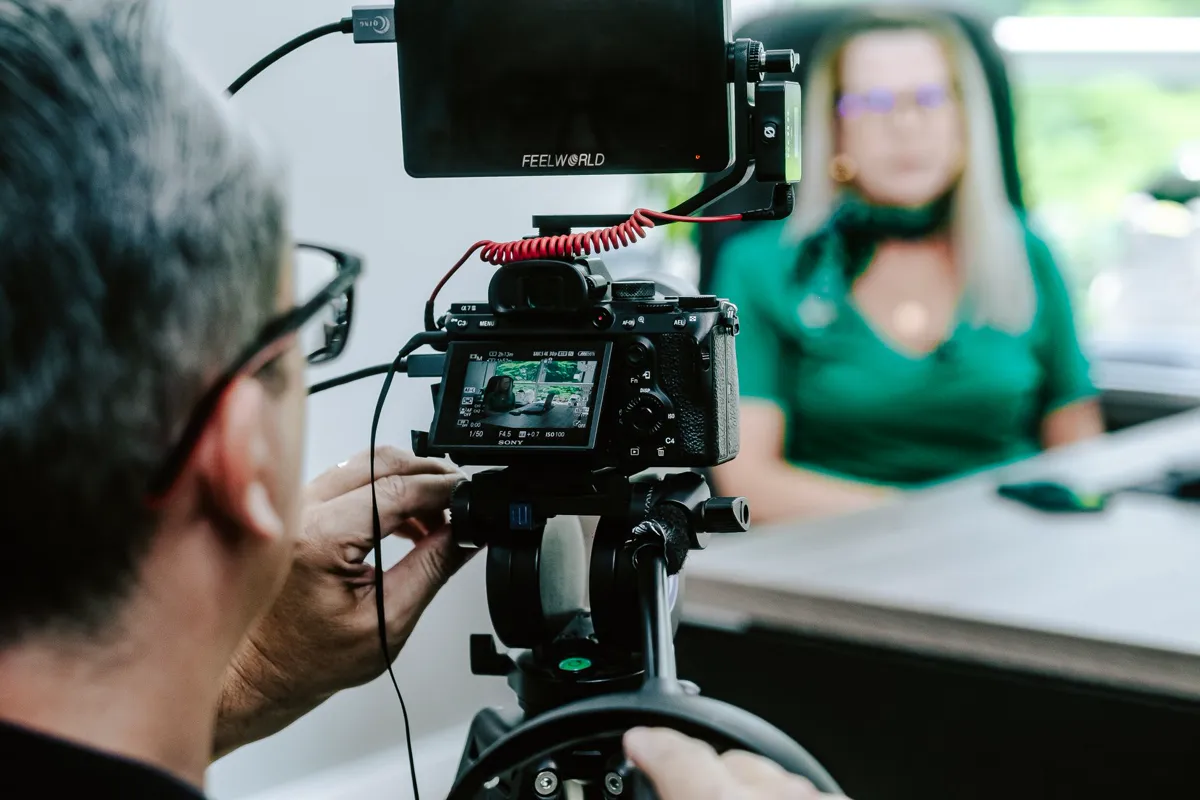 An operator behind a camera and monitor filming a seated interviewee.