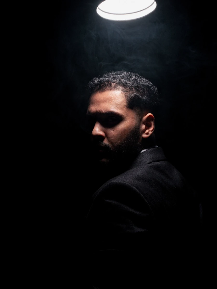 A man in a dark suit lit by a single overhead spotlight against black.