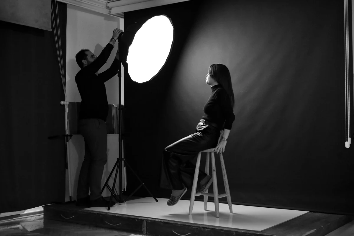 A studio shoot in progress — a crew member adjusting a large round light beside a seated model.