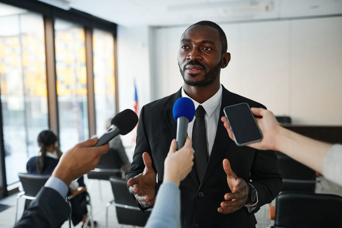 A man in a dark suit speaking to a cluster of microphones and a recording phone held out by reporters.